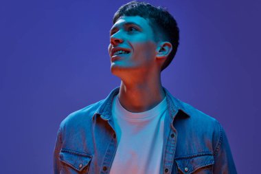 Portrait of young handsome guy in jeans shirt and white t-shirt design with smile, looking away against gradient purple background in neon light. Concept of human emotions, lifestyle, youth clipart