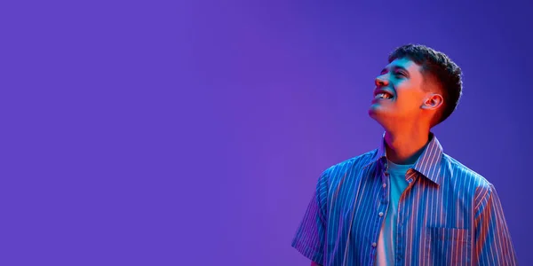 カジュアルな服を笑顔で肯定的な若い男の肖像画は ネオンの光のグラデーション紫色の背景に対して良い気分でポーズ 人間の感情 ライフスタイル 若者の概念 広告のコピースペース — ストック写真