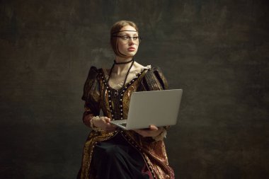 Koyu yeşil arka planda dizüstü bilgisayarla poz veren eski moda elbiseli güzel bir kızın portresi. Eğitim, serbest meslek, iş. Tarih kavramı, Rönesans sanatı, dönemin karşılaştırması.