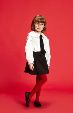 Siyah tişörtlü, beyaz bluzlu ve kırmızı arka planda duran gözlüklü küçük bir kızın tam boy portresi. Zeki, sakin, mutlu çocuk. Çocukluk, eğitim, moda, çocuk duyguları konsepti