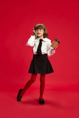 Full-length portrait of little girl in black shirt and white blouse talking on mobile phone, drinking coffee against red studio background. Concept of childhood, education, fashion, kid emotions clipart