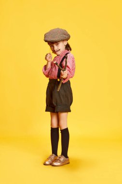 Küçük bir kızın portresi, retro giysili bir çocuk, tişört ve şort, sarı stüdyo arka planında sapanı tutuyor. Çocukluk kavramı, duygular, eğlence, moda, yaşam tarzı. Açık hava oyunu