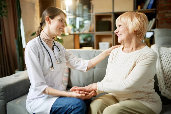 Smiling young woman, doctor supporting her elderly female patient, holding her hand and cheering up. Senior lady at home. Concept of medical care, medicine, illness, health care, profession