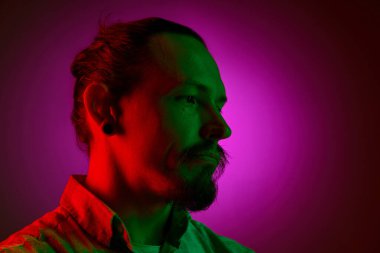 Küpeli ve at kuyruklu bir adamın yakın plan portresi sakin bir yüz ifadesiyle, neon ışıkta koyu pembe eğimli arka planda poz veriyor. İnsan duyguları, yüz ifadesi, yaşam tarzı kavramı