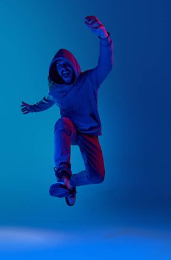 Kapüşonlu sıradan giyinmiş duygusal bir adamın uzun bir portresi neon ışıkta mavi stüdyonun arka planına zıplıyor. İnsan duyguları kavramı, yüz ifadesi, yaşam tarzı, başarı