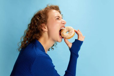 Mavi bluzlu, duygusal donut yiyen kıvırcık kızın portresi mavi stüdyo arka planında. Tatlıyı. İnsan duyguları kavramı, gençlik kültürü, moda, yaşam tarzı, yiyecek
