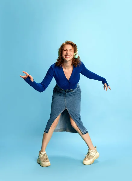 穿着蓝色上衣和牛仔裤裙的年轻快乐女孩的画像 在耳机里听音乐 在蓝色工作室的背景下在情感上跳舞 人类情感 青年文化 时尚的概念 — 图库照片