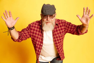 Şık, olgun, sakallı, kareli gömlekli, gözlüklü ve şapkalı bir adamın portresi. Sarı stüdyo arka planına karşı eller havada poz veriyor. İnsan duyguları kavramı, yaşam tarzı, erkek modası, tarzı