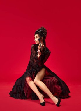 Güzel, zarif, genç bir kızın portresi makyaj yapıyor, yanaklarına kırmızı arka planda allık sürüyor. Tarih kavramı, rönesans sanatı, çağların karşılaştırması, güzellik ve moda