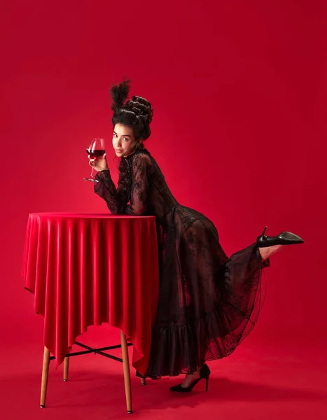 美丽迷人的年轻姑娘的画像 穿着优雅 黑色的衣服靠在桌上 手里拿着红色背景的红酒酒杯 复兴艺术 时代概念比较 — 图库照片