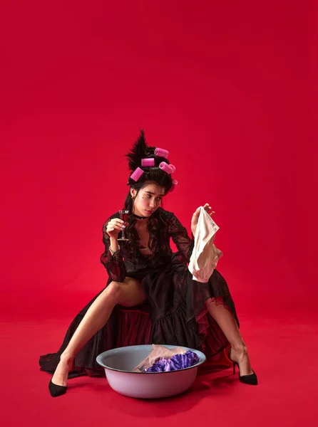 穿着卷发的年轻漂亮姑娘的画像 穿着典雅的黑色连衣裙做家务 带着枯燥无味的脸在红色的背景下洗衣服 历史的概念 复兴艺术 时代的比较 — 图库照片