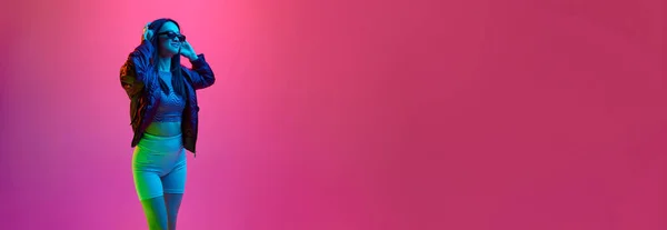 ピンクのスタジオを背景にネオンの光の中でヘッドフォンで音楽を聴く サングラスのかなり若い女の子の肖像画 感情の概念 顔の表情 ライフスタイル バナー — ストック写真