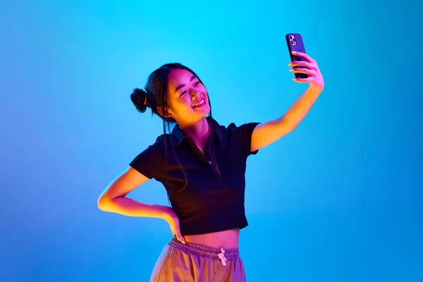在霓虹灯下 一个面带微笑的年轻的韩国女孩拿着手机在蓝色工作室背景下自拍的肖像 情感概念 面部表情 生活方式 — 图库照片