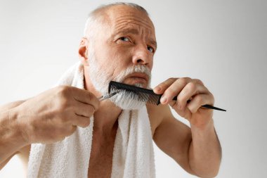 Berber. Gri saçlı, yakışıklı, olgun bir adamın portresi. Modellerin yüzüne bakması. Gri arka planda sakal kesip taraması. Erkek güzelliği, yüz ve cilt bakımı kavramı, günlük prosedürler