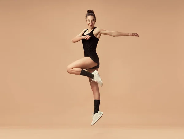 黒のスポーツウェアの訓練でスポーティな若い女の子のダイナミックなイメージは 明るい茶色のスタジオの背景にジャンプします 筋肉を作る スポーツライフスタイル 美しさ ボディケア フィットネス 健康の概念 — ストック写真