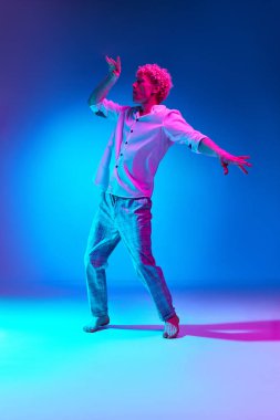 Beyaz gömlekli ve kareli pantolonlu olgun bir adamın pembe neon ışıkta mavi stüdyo arka planında dans edişinin tam boy portresi. İnsan duyguları kavramı, yaşam tarzı, gençlik, yüz ifadesi