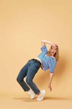 Sarı stüdyo arka planında poz veren çizgili tişört ve kot giyen sarışın bir kızın tam boy portresi. Öğrenci. Gençlik kavramı, insan duyguları, yüz ifadesi