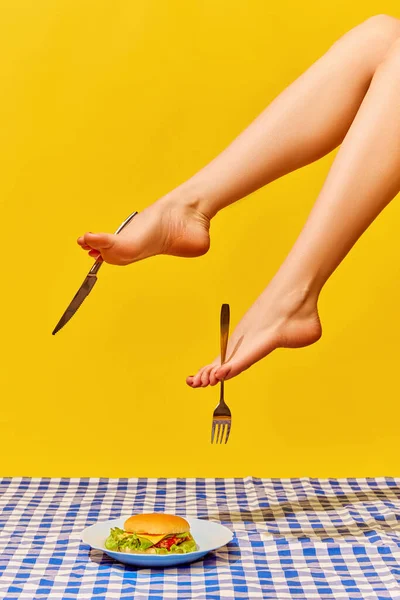 女性细长腿拿着叉子和刀 在鲜活的黄色背景下吃着美味的汉堡包的创造性形象 流行艺术摄影 创意视觉 想象力的概念 最低限度艺术 — 图库照片