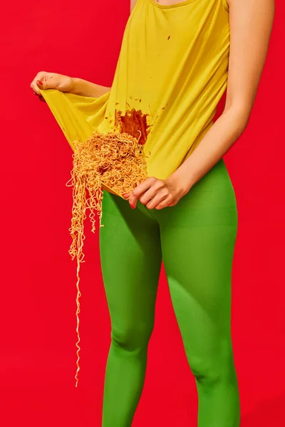 Cropped image of female legs in green tights and yellow t-shirt with noodles inside against vivid red background. Concept of pop art photography, creative vision, imagination. Complementary colors
