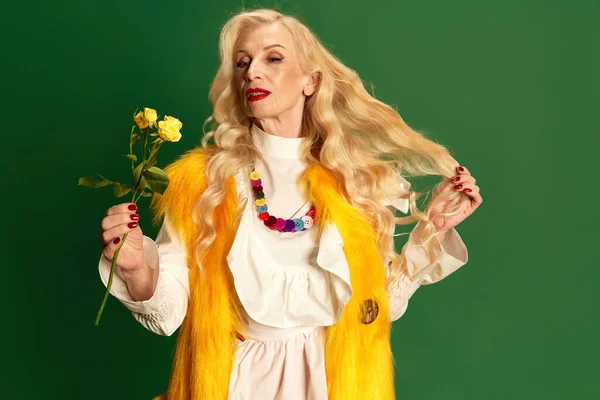 stock image Portrait of beautiful lady with long blonde hair and makeup, senior woman in stylish clothes holding yellow roses against green studio background. Concept of beauty, fashion, human emotions, lifestyle