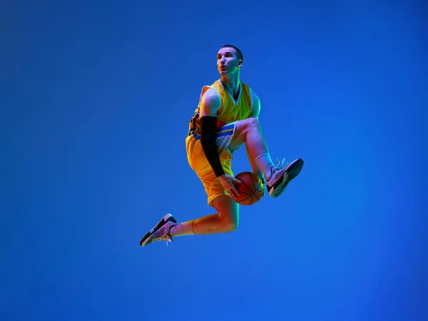 ネオンの光の中で青いスタジオの背景にボールでジャンプし 動きのプロの男性バスケットボール選手のダイナミックなイメージ プロスポーツの概念 健康的なライフスタイル アクションと — ストック写真