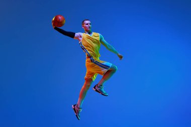 Sarı üniformalı kaslı genç bir adam, maç sırasında basketbol oyuncusu, neon ışıkta mavi stüdyonun arka planında topla zıplıyor. Profesyonel spor, hobi, sağlıklı yaşam tarzı, eylem kavramı