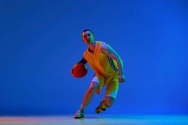 Maç sırasında sarı üniformalı erkek basketbolcu, neon ışıkta mavi stüdyonun arka planına top sürüyor. Profesyonel spor, hobi, sağlıklı yaşam tarzı, aksiyon ve hareket kavramı, oyun