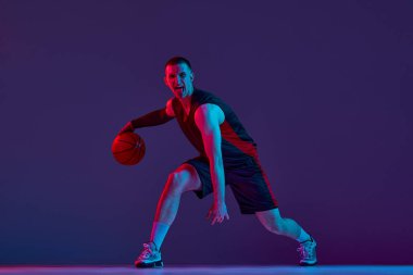 Genç kaslı bir adam, hareketli bir basketbol oyuncusu, neon ışıkta mor stüdyonun arka planına top sürüyor. Kazandım. Profesyonel spor, hobi, sağlıklı yaşam tarzı, eylem ve hareket kavramı