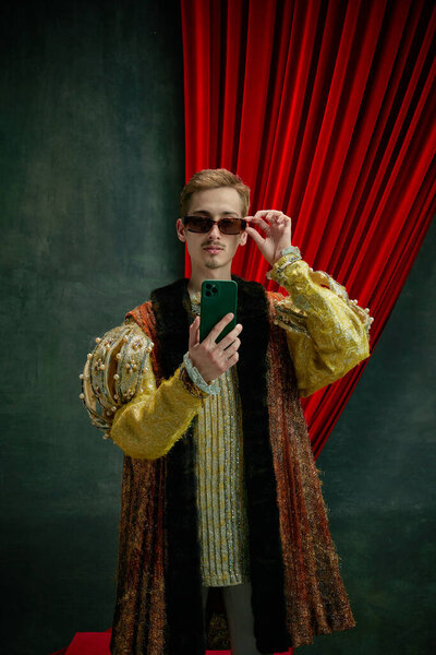 Portrait of young man in stylish clothes and sunglasses, royal person checkign social media against dark green, vintage background. Concept of comparison of eras, history, renaissance art, remake