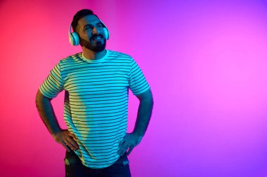 Çizgili tişörtlü sakallı bir adamın portresi kulaklıkla müzik dinliyor. Neon ışıklarıyla mavi renkli gradyan stüdyonun arka planına karşı. İnsan duyguları, yüz ifadesi, yaşam tarzı kavramı