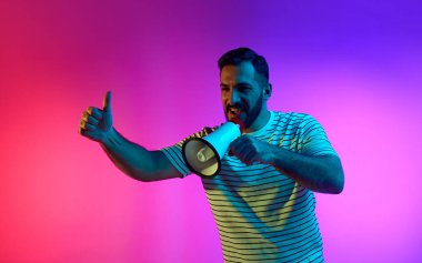 Sakallı, mutlu bir adam megafonla konuşuyor ve neon ışıklarıyla pembe renkli stüdyo arka planına karşı elleri ile olumlu bir jest yapıyor. İnsan duyguları, yüz ifadesi, yaşam tarzı kavramı