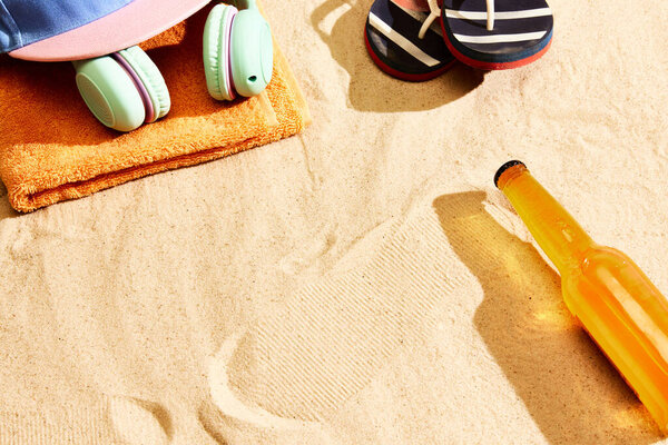 Bottle of lager beer on warm sand with slippers, headphones, towel and cap. Summertime relaxation. Concept of alcohol drink, taste, summer vacation, holiday, brewery. Advertisement