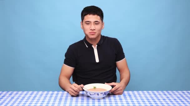 青いスタジオの背景に麺でスパイシーなスープを食べる顔を食いしばるハンサムな若い男 スパイシーなホット料理スタイル 味の概念 人間の感情 顔の表情 — ストック動画