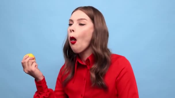 青いスタジオの背景にレモンをかむ若い美しい女の子 甘酸っぱい柑橘類を食べる顔を食いしばる感情的な女性 味の概念 人間の感情 顔の表情 — ストック動画