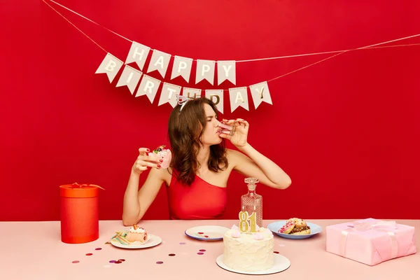 Getting older, feeling sad and depressed. Young birthday girl drinking alcohol, celebrating alone against red background. Concept of party, celebration, emotions, female beauty, youth. Pop art