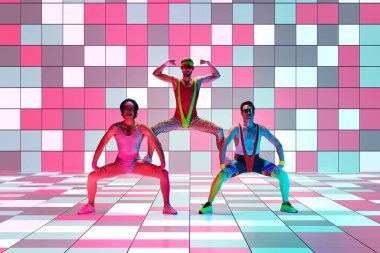 Üç adam şık, klasik spor giyim eğitiminde, neon ışıkta mozaik stüdyosunun arka planında aerobik egzersizleri yapıyor. Sportif ve aktif yaşam tarzı, mizah, eski tarz kavramı. Ad