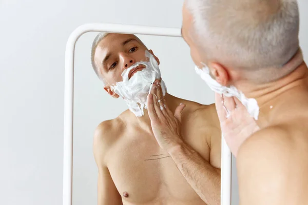白いスタジオの背景に鏡を見て 彼のひげを剃って若い恥知らずの男の肖像画 男性の美しさ スキンケア 健康の概念 広告のコピースペース — ストック写真