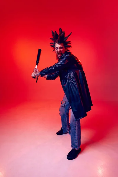Expressive young man, punk with weird hairstyle and makeup, aggressively posing with baseball bat over red studio background in neon light. Music, lifestyle, subculture, art, youth, emotions concept