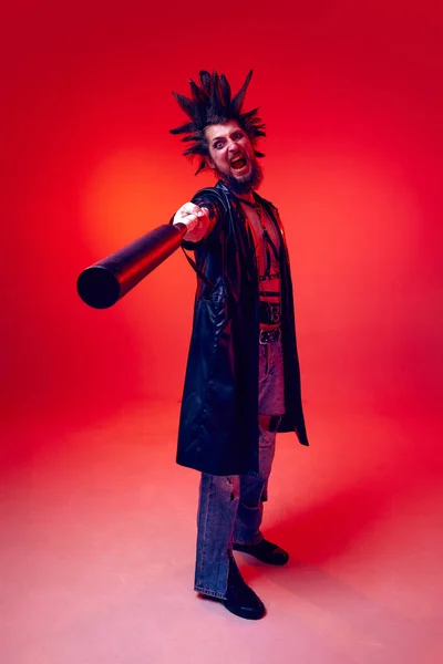 Expressive young man, punk with weird hairstyle and makeup, aggressively posing with baseball bat over red studio background in neon light. Music, lifestyle, subculture, art, youth, emotions concept