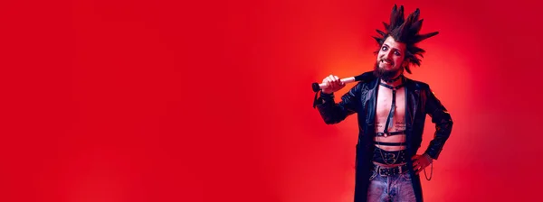 Crazy looking emotive young man, punk with expressive hairdo posing with bat over red background in neon light. Concept of music, lifestyle, subculture, art, youth, emotions. Banner. Copy space for ad