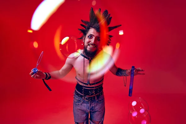 Crazy looking young man, punk, musician posing shirtless with soap bubbles against red studio background in neon light. Concept of music, lifestyle, subculture, art, youth, human emotions