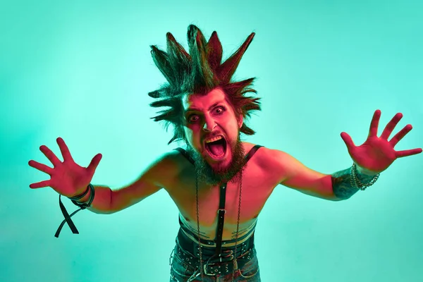 Portrait of young crazy looking man, punk with expressive hairstyle posing shirtless against blue studio background in neon light. Concept of music, lifestyle, subculture, art, youth, human emotions