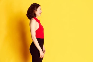 Sıradan giyinen, poz veren, sarı stüdyo geçmişine bakan esmer bir kadının profil görüntüsü. Gençlik kavramı, insan duyguları, yaşam tarzı, güzellik, moda, reklam