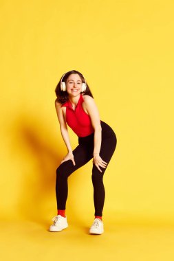 Gündelik giysiler içinde, kulaklıkla müzik dinleyen ve sarı stüdyo arka planında dans eden genç güzel bir kadının tam boy portresi. Gençlik kavramı, insani duygular, yaşam tarzı, reklam