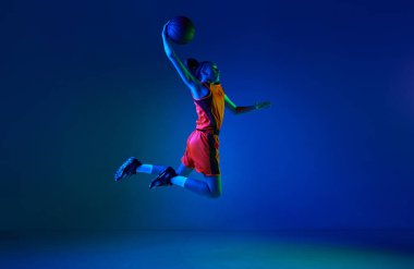 Smaç basıldı. Motive olmuş genç bir kız, basketbolcu neon ışıkta mavi stüdyonun arka planına karşı sıçrıyor. Profesyonel spor, eylem ve hareket kavramı, oyun, rekabet, hobi, reklam