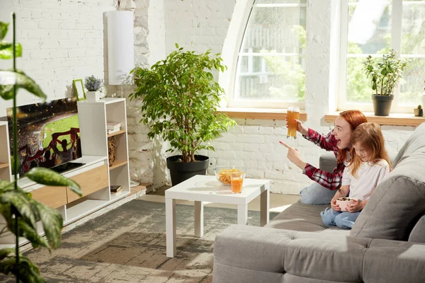 stock image Young woman sitting on couch at home, drinking beer, watching football match on tv with little bored girl, child eating cereal. Concept of hobby, entertainment, lifestyle, sport, competition, emotions