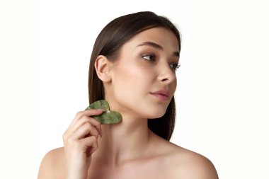 Güzel, sağlıklı cilde sahip genç bir kadın beyaz arka plana karşı kozmetik masaj aleti ile yüzüne dikkat ediyor. Doğal kadın güzelliği kavramı, vücut ve cilt bakımı, kozmetoloji, reklam