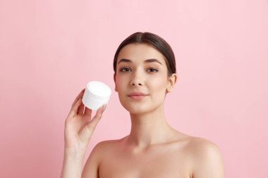 Pembe stüdyo arka planında yüzü nemlendirici kremle poz veren güzel bir kadının portresi. Organik kozmetik ürünleri. Doğal kadın güzelliği kavramı, vücut ve cilt bakımı, kozmetoloji, sağlık, reklam
