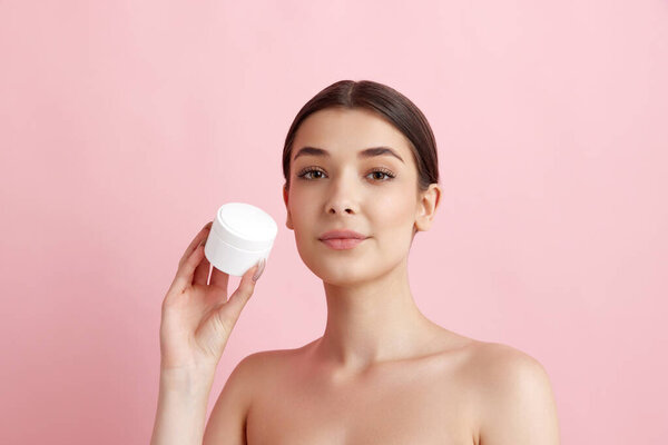 Portrait of young beautiful woman posing with face moisturizing cream against pink studio background. Organic cosmetics. Concept of natural female beauty, body and skincare, cosmetology, health, ad