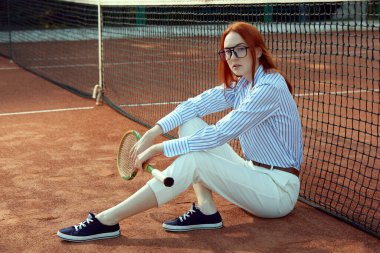 Beyaz pantolonlu, çizgili tişörtlü, şık gözlüklü, tenis kortunda tenis raketiyle poz veren güzel kızıl saçlı kadın. Spor, aktif yaşam tarzı, günlük moda, boş zaman, reklam konsepti.